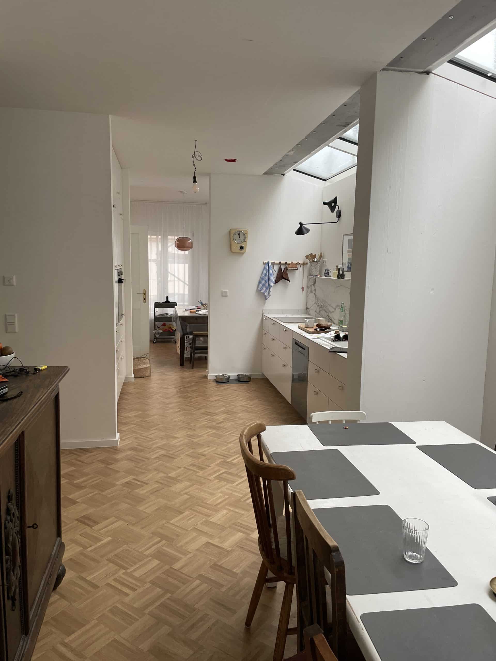 Vordergrund: Essplatz mit offenem Durchgang zur Küche mit einer Küchenzeile rechts, Hintergrund: Arbeitszimmer; Boden: Parkett