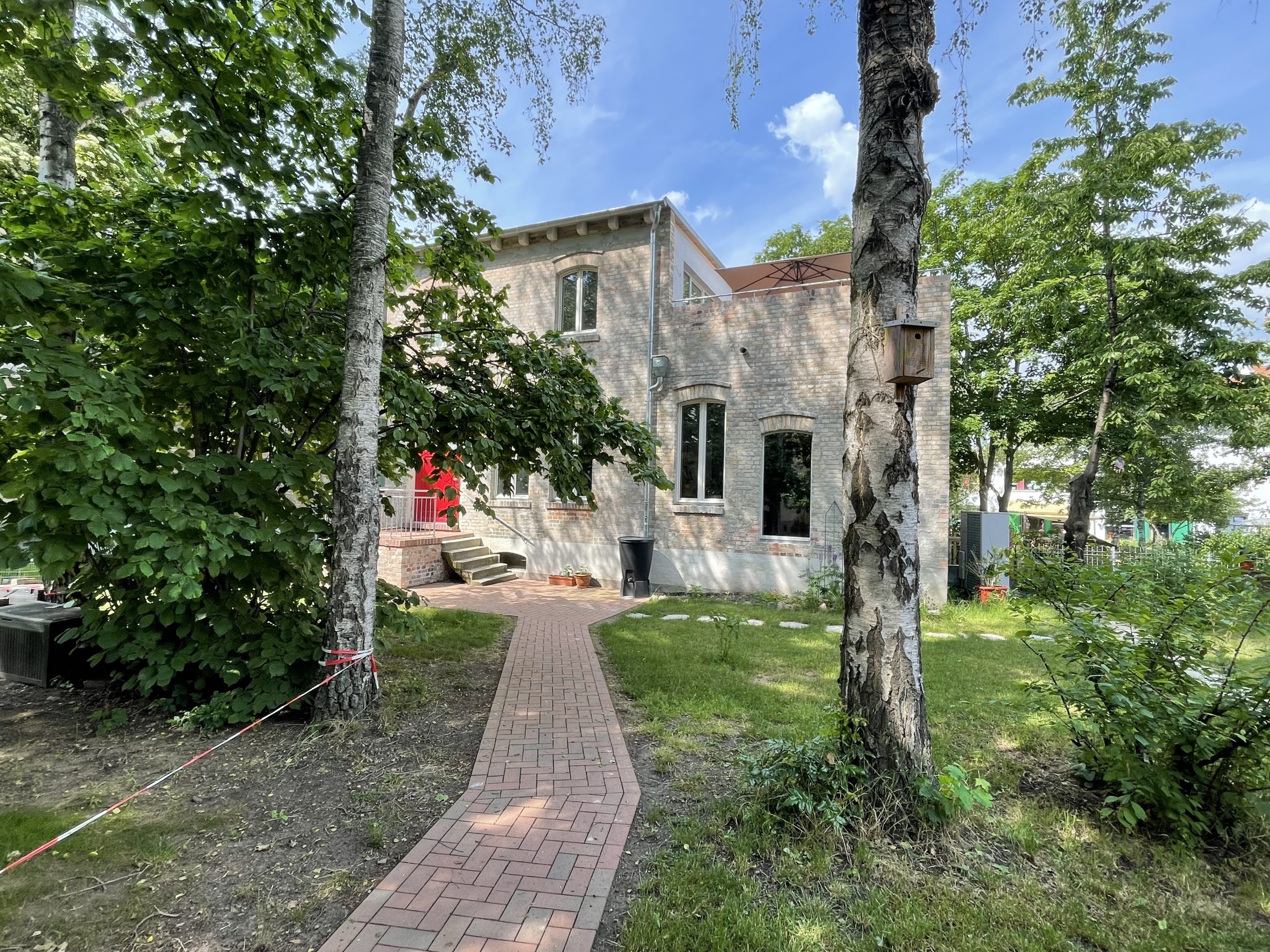 Fotografie des Hinterhauses Glauchaviertel mit dem Weg zum Haus durch den Garten und zwei Bäumen im Vordergrund und einem Baum rechts neben dem Haus im Hintergrund.