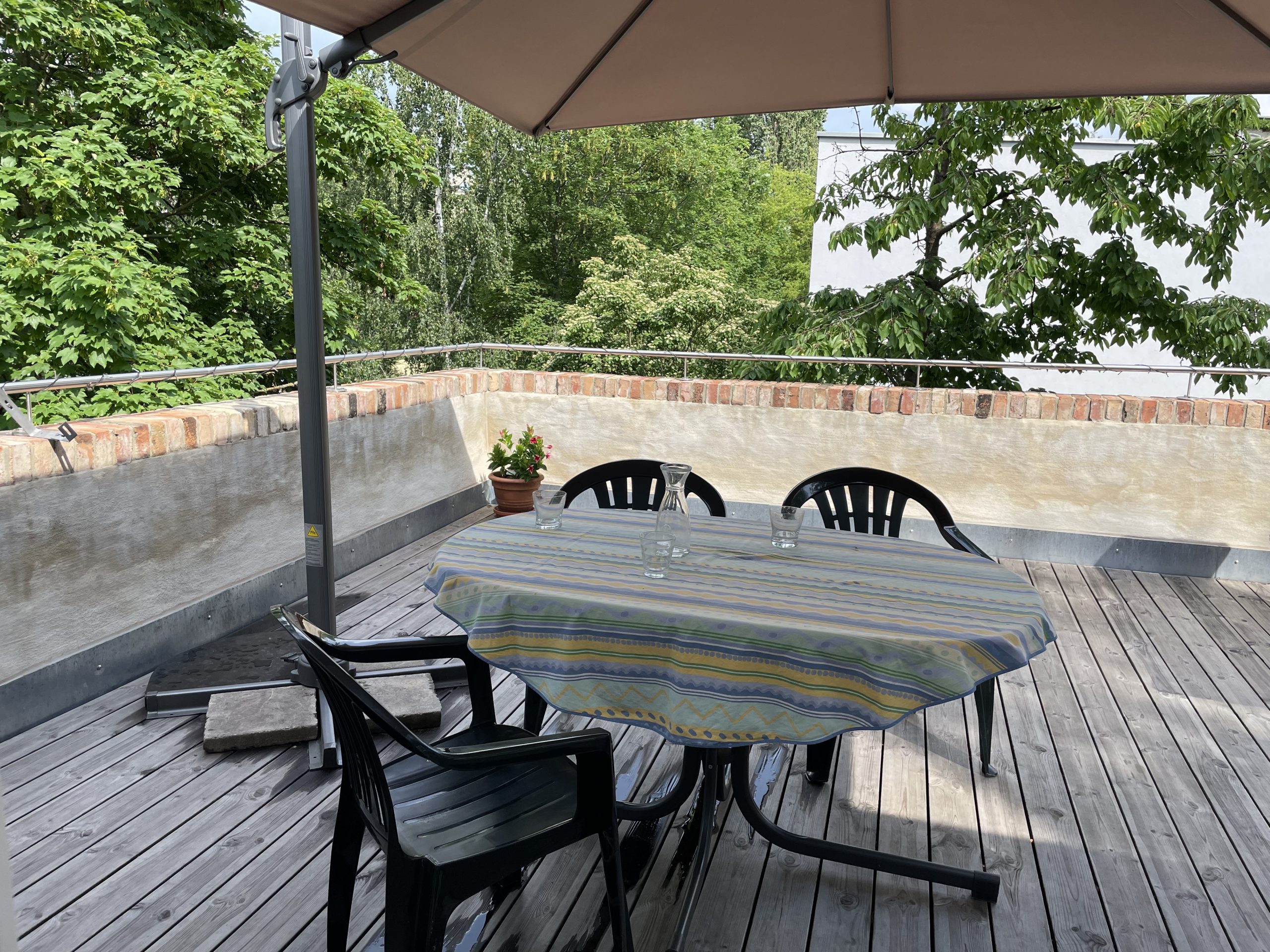 Fotografie der Terrasse des Hinterhauses Glauchaviertel mit Holzboden, auf dem ein Esstisch, drei Stühle und ein Sonnenschirm stehen, hinter der Terrasse befinden sich Bäume.